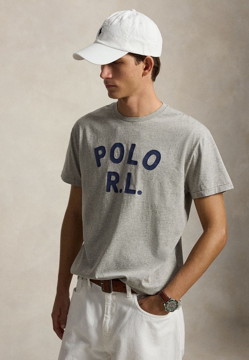 Νέος άνδρας που φοράει γκρι μπλούζα "POLO R.L.", άσπρο καπέλο, άσπρο παντελόνι, καφέ ζώνη και ρολόι χειρός, στέκεται μπροστά σε ουδέτερο φόντο.