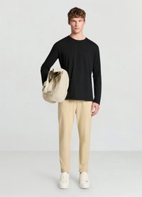 Schwarzes Langarmshirt, beige Hose und weiße Sneaker. Die Person hält eine weiche beige Tasche mit zwei Griffen, die ein minimalistisches Design aufweist.