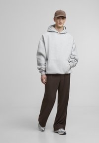Grauer Hoodie mit einer frontalen Tasche, kombiniert mit braunen, weit geschnittenen Hosen und silbernen Turnschuhen. Eine braune Kappe wird getragen, die mit gesticktem Text verziert ist.