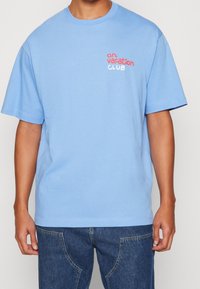 Ljusblå bomullströja, kort ärm, rundad halsringning. Har texten "on vacation" i röd och "CLUB" i vit broderad text på vänster bröst.