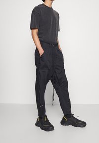 Schwarzes Baumwoll-T-Shirt, lockere Passform. Schwarze Cargo-Hose mit Kordelzugbund, mehreren Taschen und sichtbaren Nähten. Schwarze Sneaker mit gelben Akzenten.