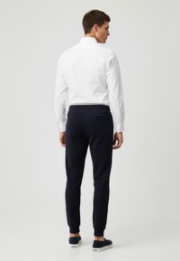 Weißes Langarmhemd mit Knopfmanschetten, getragen mit marineblauen, schmal geschnittenen Jogginghosen mit elastischem Bund und dezentem TaschenDetail auf der Rückseite.