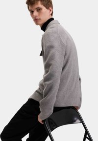 Szary sweter dzianinowy o prążkowanej fakturze, z kołnierzem i detalami w postaci guzików po bokach, zestawiony z czarnymi spodniami, siedzący na czarnym krześle.