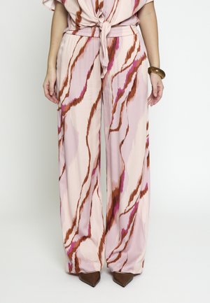 Donna che indossa pantaloni rosa a gamba larga con strisce astratte marroni e viola, abbinati a scarpe marroni con punta e braccialetto di legno.