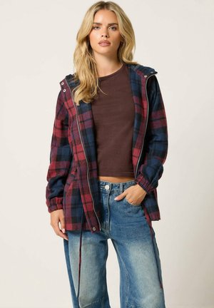 Femme aux cheveux blonds portant une veste zippée à carreaux rouge et bleu marine, un t-shirt court marron et un jean bleu délavé avec une main dans la poche.