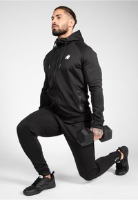 L'outfit sportivo nero include un hoodie con zip, tasche laterali e un logo, pantaloni jogger abbinati e scarpe da allenamento nere. Tiene dei manubri.