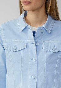 Veste en denim bleu clair avec deux poches poitrine, col angulaire et fermeture à boutons. Texture lisse avec de subtils détails de couture.