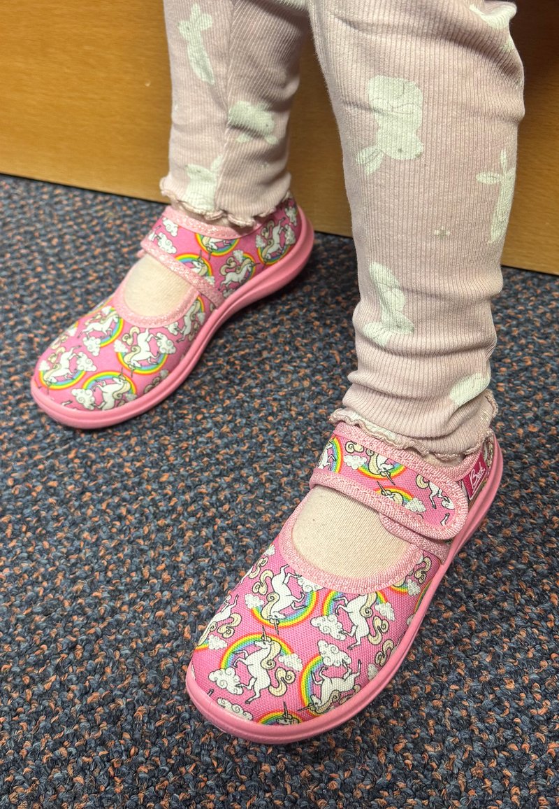Kind trägt rosa Schuhe mit Einhorn- und Regenbogenmuster, beige Socken und blassrosa Leggings mit weißen Hasenmotiven und steht auf einem Teppichboden.