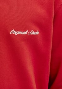 Felpa rossa realizzata in morbido tessuto, con il testo ricamato "Originals Studio" in bianco sul lato sinistro superiore.