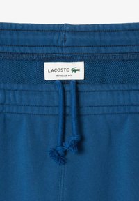 Cintura de algodón azul con cordones ajustables, que presenta una etiqueta de logo de Lacoste que dice "FIT NORMAL". Textura suave y color sólido.