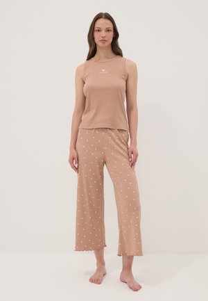 Woman standing barefoot wearing beige sleeveless top and wide-leg pants with small white heart patterns on a plain background.