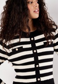 Cardigan a strisce bianche e nere, con texture a costine, dotato di bottoni perlate e due tasche sul petto con dettagli neri.