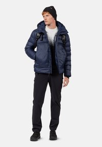 Giacca piumino blu marina con cappuccio, abbinata a un berretto nero, maglione monocromo, pantaloni scuri e scarpe nere. Include spallacci.
