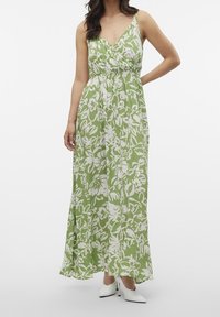 Žena v zeleno-bílých květovaných bezramínkových maxi šatech s bílými špičatými vysokými podpatky, stojící s jednou rukou na boku.
