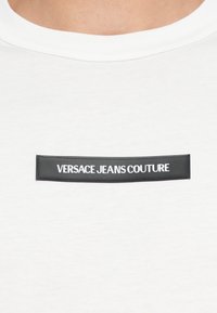 Hvit bomullst-skjorte med en svart rektangulær lapp med "VERSACE JEANS COUTURE" i hvit skrift, plassert i midten.