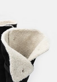 Czarne buty z miękkim, kremowym ociepleniem z polaru, złożonym na górze, ozdobione srebrnymi elementami oraz teksturowanym zewnętrznym materiałem.