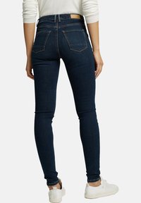 Jeans ajustados en denim azul oscuro, con cintura alta, dos bolsillos traseros y detalles de costura. Llevados con zapatillas blancas.