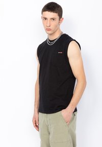 Maglietta senza maniche nera con taschino, realizzata in cotone, indossata con pantaloni cargo verde chiaro, accessoriata con una collana in argento.