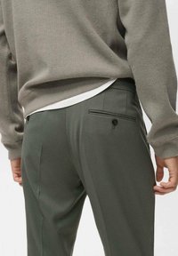 Pantalones de corte oscuro en verde, con una textura suave, que presentan un bolsillo trasero y un detalle de botón. Combinados con un suéter de punto gris claro.