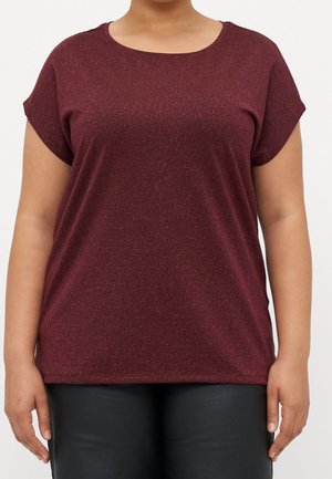 Person trägt ein kurzärmliges, funkelndes burgunderrotes Oberteil und schwarze Hosen, steht vor einem schlichten hellen Hintergrund.