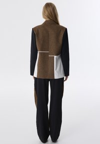 Bruine en zwarte getailleerde blazer met een gestructureerde achterkant, gecombineerd met brede zwarte broek met bruine accenten en een gestreept detail op de taille.