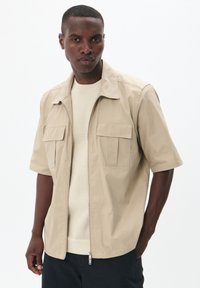 Jachetă bej cu mânecă scurtă, cu fermoar în față, două buzunare pe piept și o textură ușoară și netedă, purtată peste un tricou din tricot de culoare crem.