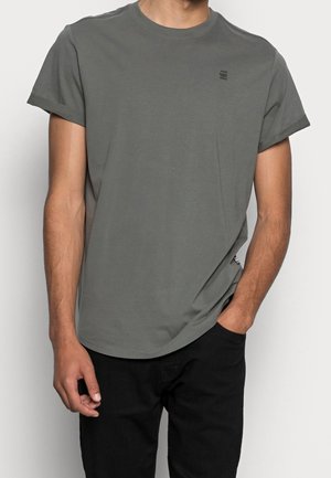 Mężczyzna ubrany w gładki szary t-shirt z krótkim rękawem i czarne spodnie, z jedną ręką w kieszeni, a drugą swobodnie opuszczoną wzdłuż ciała.