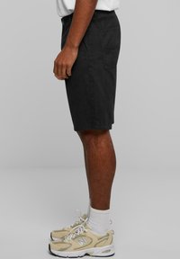 Pantalonii scurți din bumbac negri au un croi drept, două buzunare laterale și un buzunar pe spate. Sunt purtați cu adidași bej și șosete albe.