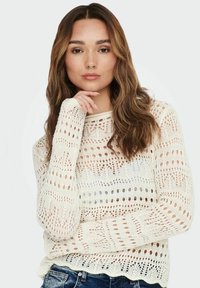 Giovane donna con capelli lunghi e mossi che indossa un maglione bianco a maglia aperta e jeans blu, poggia il mento sulla mano, sfondo neutro.