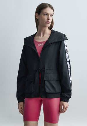 Femme portant une veste zippée noire avec des manches blanches à logo, par-dessus un haut de sport rose et un cuissard vélo assorti, debout sur un fond uni.