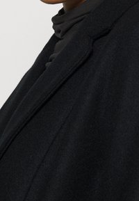 Manteau en laine noire avec un col à revers cranté, des épaules structurées et une texture douce. Gros plan sur la zone du col et le tissu adjacent.