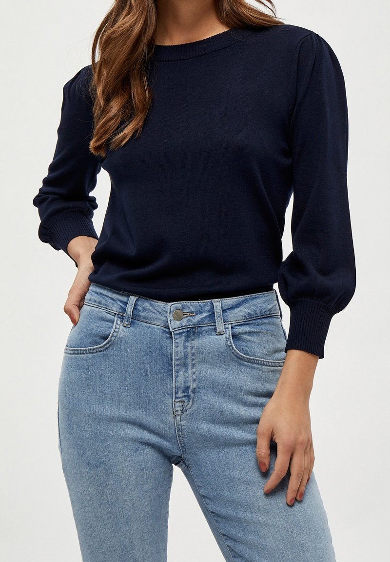 Modell trägt einen marineblauen Pullover und hellblaue Jeans, steht mit einer Hand auf der Hüfte vor einem hellgrauen Hintergrund.