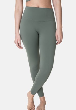 Frau trägt hoch taillierte, olivgrüne Leggings, steht barfuß mit gekreuzten Beinen vor einem einfarbigen Hintergrund.