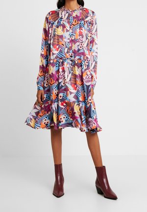 Mujer vestida con un vestido floral colorido de manga larga y largo hasta la rodilla, combinado con botines burdeos, de pie frente a un fondo liso.