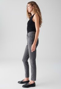 Donna con lunghi capelli ricci, che indossa una canotta nera senza maniche, jeans skinny grigi e mocassini neri, in profilo su uno sfondo semplice.