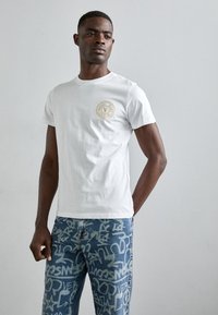 Λευκό βαμβακερό μπλουζάκι με στρογγυλή λαιμόκοψη και κυκλικό λογότυπο σε χρυσό στο αριστερό στήθος. Φοριέται με μπλε τζιν που εμφανίζουν μοτίβο γκράφιτι.