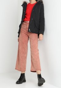 Svart parkas med foder av päls i huvan, röd tröja och högmidjade ljusrosa vida jeans. Svarta snörstövlar vid anklarna fulländar outfiten.