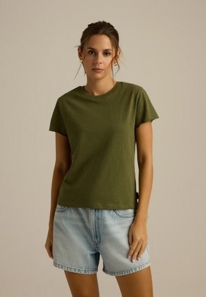 Oliwkowa koszulka z krótkim rękawem, wykonana z miękkiego materiału, luźny fason, okrągły dekolt, w połączeniu z jasnoniebieskimi szortami dżinsowymi.