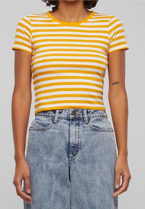 Vrouw draagt een geel-wit gestreept cropped shirt met korte mouwen en een lichtblauwe high-waisted spijkerbroek tegen een effen grijze achtergrond.