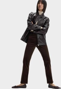 Schwarze Kunstlederjacke, beiger Rollkragenpullover, dunkelbraune Cordhosen und schwarze Ballerinas. Das Model steht selbstbewusst mit verschränkten Armen.