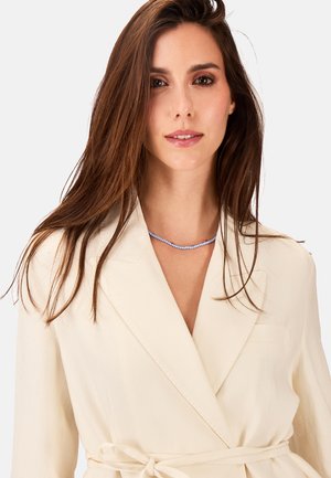Cremfarbener Wickelblazer aus strukturiertem Stoff mit Taillenknoten. Eine Halskette aus blauen und roten Perlen betont den Ausschnitt.