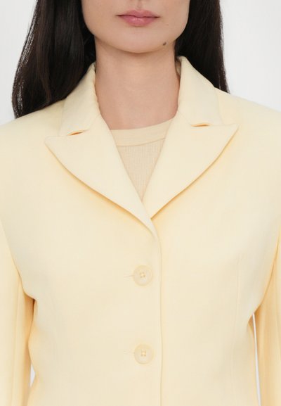 Light yellow blazer with a notched collar, three front buttons, and a smooth texture. Worn over a cream-colored top with ribbed detail.