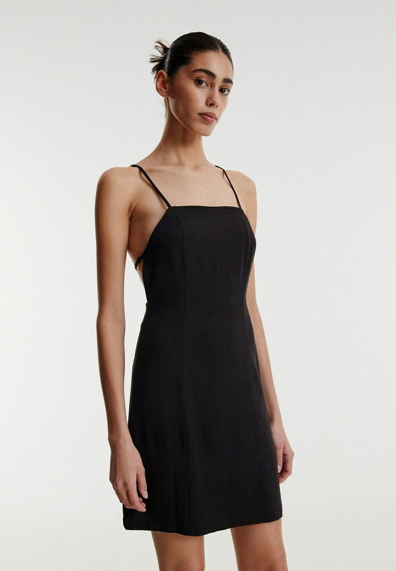 Schwarzes Kleid aus leichtem Stoff, mit einem eng anliegenden Oberteil, dünnen verstellbaren Trägern und einer geraden Silhouette, die oberhalb des Knies endet.