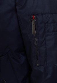 Tmavě námořnická bunda s kapsou a vertikálním kovovým zipem a červeným textilním detailem na základně zipu.