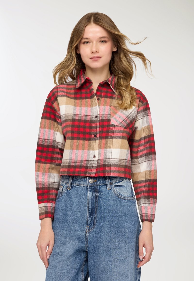 Camicia a quadri corta in rosso, beige e marrone con bottoni frontali e una tasca sul petto, abbinata a jeans di denim blu a vita alta.