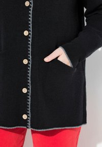 Personne portant un cardigan noir à boutons en bois et bord gris, une main dans la poche, associé à un pantalon rouge vif.