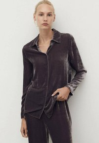 Camisa de terciopelo de manga larga en gris oscuro con collar, cierre de botones y textura suave. Presenta un corte relajado y bolsillos laterales.