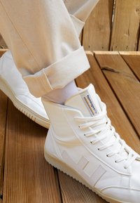Białe skórzane sneakersy za kostkę z teksturowanym wzorem, płaskimi sznurowadłami i wielokolorowym logo z boku. Jasne spodnie z podwiniętymi nogawkami.