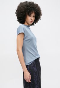 Donna con capelli ricci che indossa una maglietta a maniche corte azzurro chiaro e una gonna scura con motivo in pelle di serpente viola, in piedi di profilo su sfondo bianco.