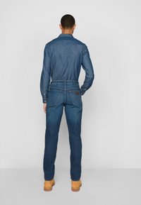 Chemise en denim bleu foncé à manches longues et col, associée à un pantalon en denim assorti. La vue arrière montre des coutures détaillées et une étiquette en cuir.